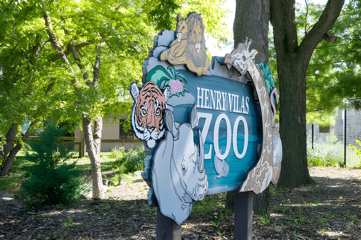 Entrance sign for the Henry Vilas Zoo in Madison, WI. Colorful wood sign with animals. Trees and landscaping in the background.